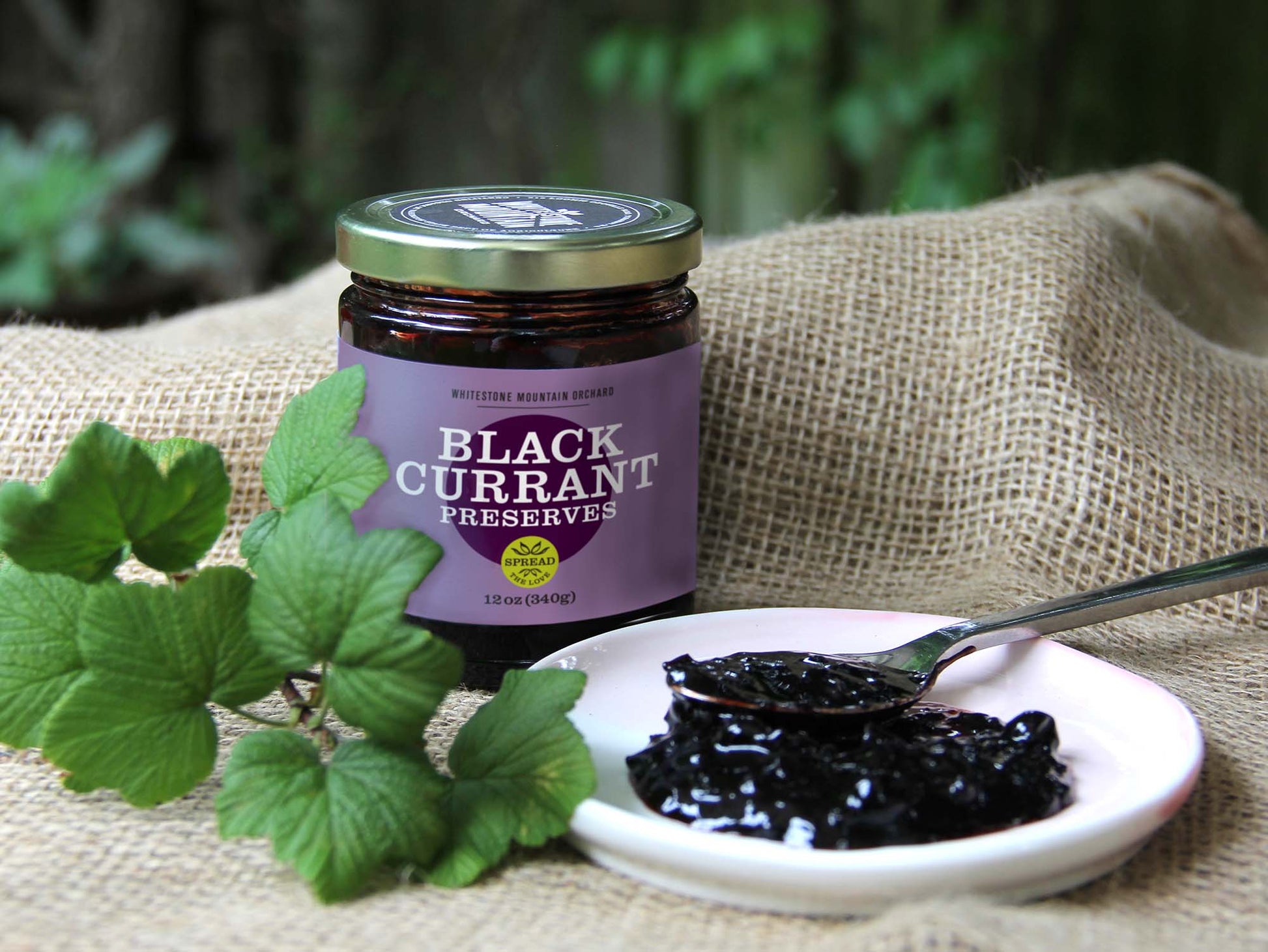A 12oz jar of Black Currant Preserves from Whitestone Mountain Orchard sits on a brown burlap fabric, with rich, dark preserves spooned onto a small plate next to it.