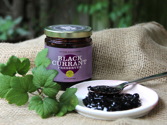 A 12oz jar of Black Currant Preserves from Whitestone Mountain Orchard sits on a brown burlap fabric, with rich, dark preserves spooned onto a small plate next to it.