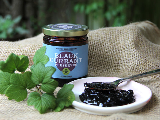 A 12oz jar of black currant preserves from Whitestone Mountain Orchard, next to a plate with deep purple preserves spooned onto it.